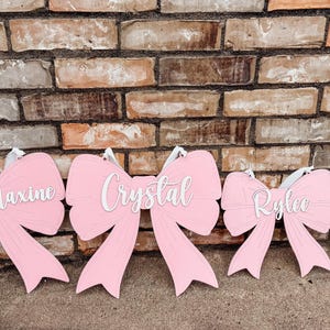 May include: Five pink wooden bows with white lettering. The bows are decorated with the names Ada Maxine, Crystal, Rylee, and Landry.