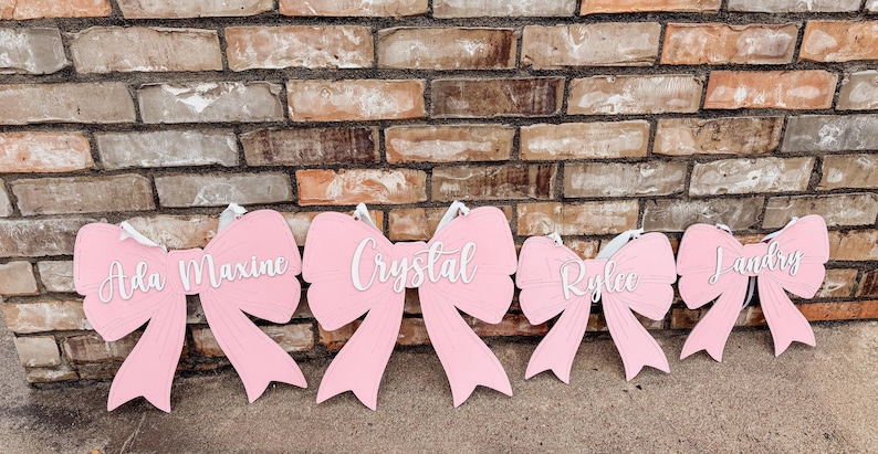 May include: Five pink wooden bows with white lettering. The bows are decorated with the names Ada Maxine, Crystal, Rylee, and Landry.