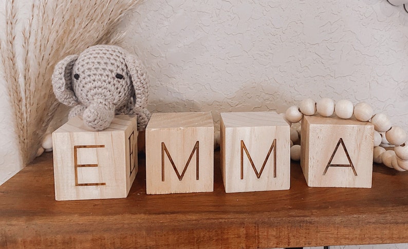 Puede incluir: Bloques de madera que deletrean el nombre "EMMA" en may&uacute;sculas. Los bloques son de color marr&oacute;n claro y tienen una veta de madera natural. Un peque&ntilde;o elefante de crochet gris se sienta encima del primer bloque.