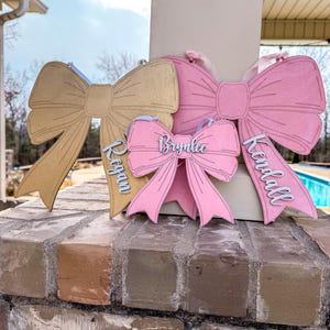 May include: Three wooden bow-shaped signs with names written on them. The top bow is gold, the middle bow is pink, and the bottom bow is pink. The names on the signs are "Brynnlee", "Kendall", and "Reagan".