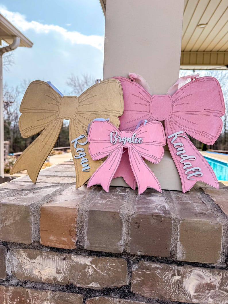May include: Three wooden bow-shaped signs with names written on them. The top bow is gold, the middle bow is pink, and the bottom bow is pink. The names on the signs are "Brynnlee", "Kendall", and "Reagan".
