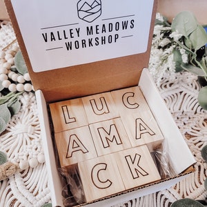 Puede incluir: Una caja blanca con el texto "Valley Meadows Workshop" en la tapa. La caja est&aacute; abierta y contiene bloques de madera con las letras "LUCAMCK" deletreadas.