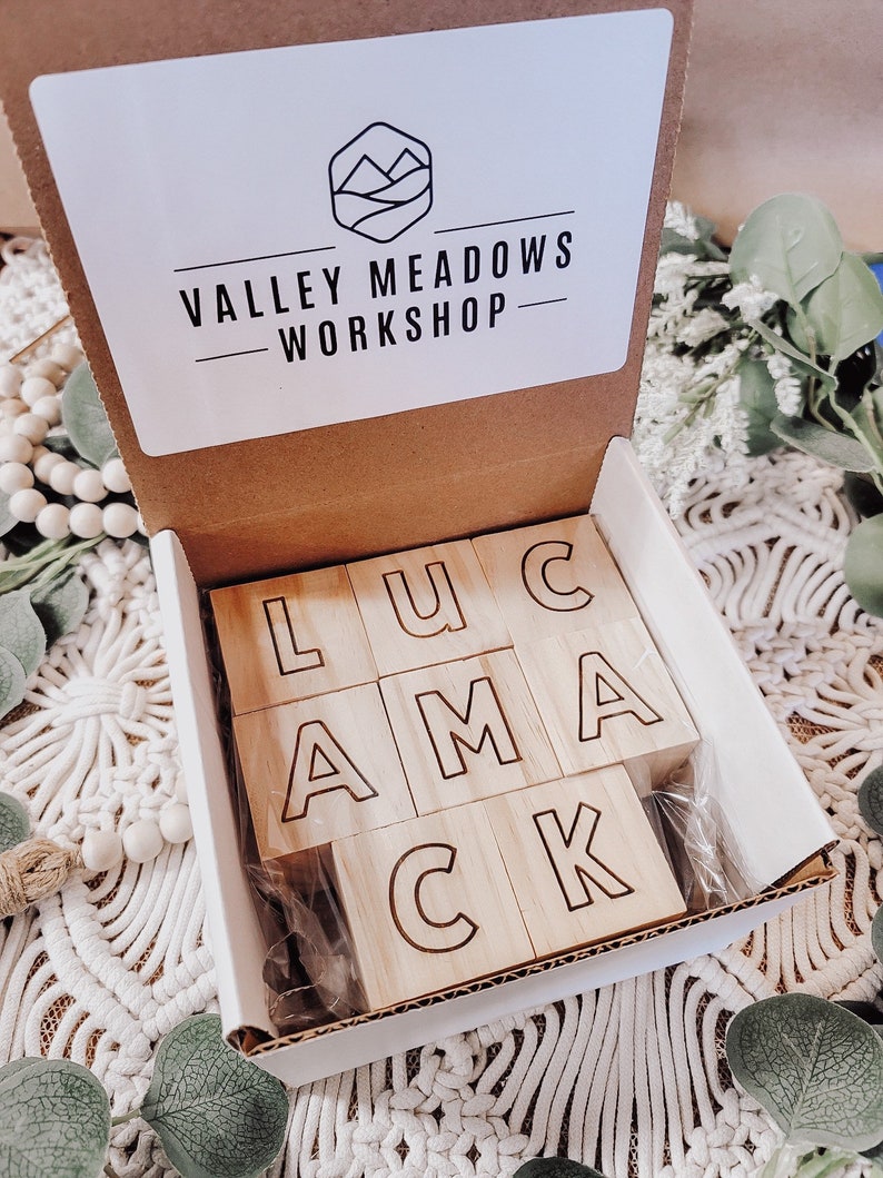 Puede incluir: Una caja blanca con el texto "Valley Meadows Workshop" en la tapa. La caja est&aacute; abierta y contiene bloques de madera con las letras "LUCAMCK" deletreadas.