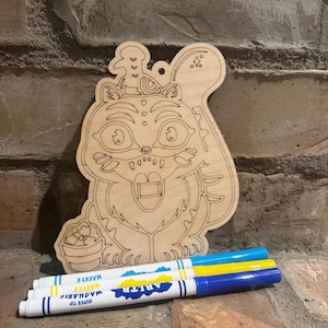 May include: A wooden cat-shaped craft with a bird on its head, a basket of food, and a rectangular opening. Three colorful markers are placed below the wooden craft. The markers are white with blue, yellow, and blue caps.