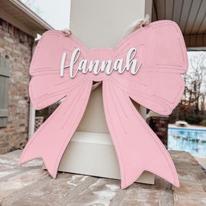 May include: A pink wooden bow with the name "Hannah" written in white on the centre. The bow is hanging from a white string.