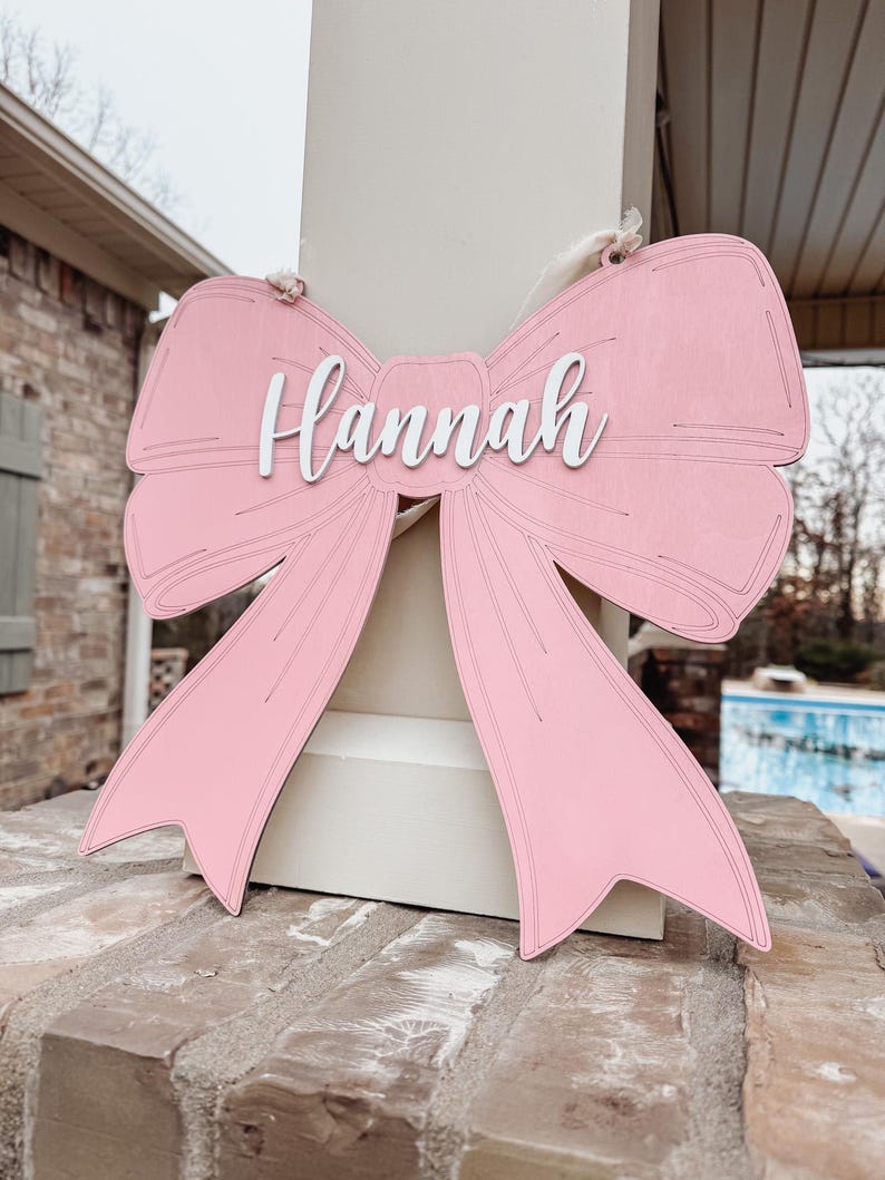 May include: A pink wooden bow with the name "Hannah" written in white on the centre. The bow is hanging from a white string.