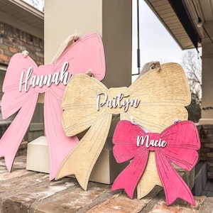 May include: Three wooden bow-shaped signs with the names "Hannah", "Paityn", and "Macie" written in white lettering. The bows are painted in pink, light brown, and red.