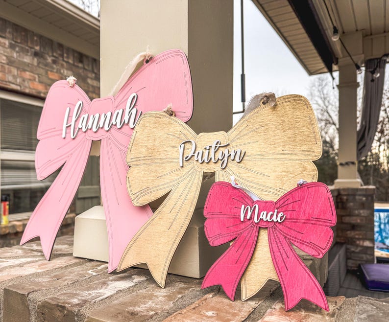 May include: Three wooden bow-shaped signs with the names "Hannah", "Paityn", and "Macie" written in white lettering. The bows are painted in pink, light brown, and red.