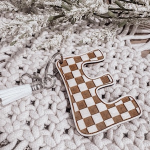 May include: A wooden keychain with a checkered pattern in the shape of the letter "E". The keychain has a white tassel attached to a silver key ring.