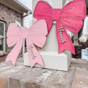 May include: Two pink wooden bows with the name "Hannah" written on them. The bows are hanging on a white post.