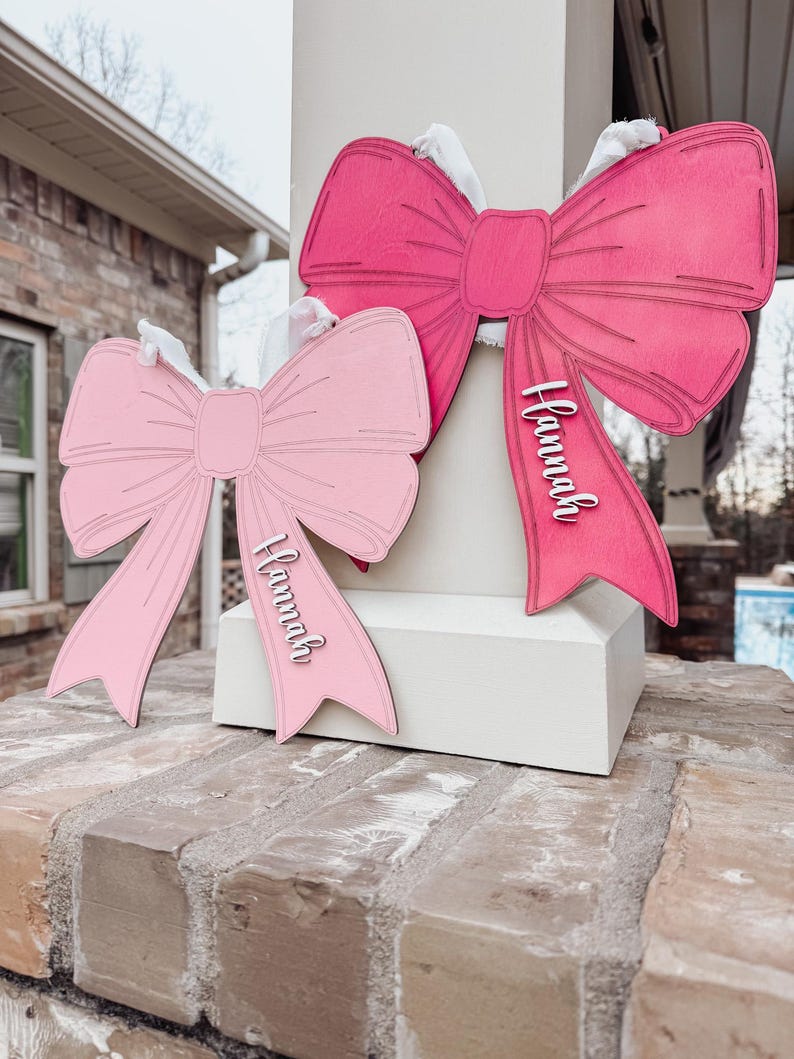 May include: Two pink wooden bows with the name "Hannah" written on them. The bows are hanging on a white post.