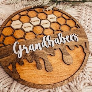 May include: A wooden honeybee-themed sign with the words "Grandbabes" and the names "William", "Brody", "Paisley", "Lainey", and "Mila" inscribed in the honeycomb design.