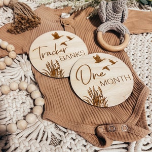May include: Two wooden baby milestone discs with a nature theme. The first disc says "Tracker Banks" with a design of birds and reeds. The second disc says "One Month" with a similar design.