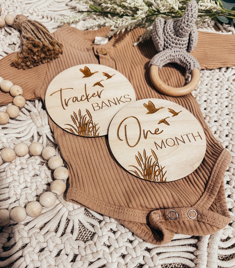 May include: Two wooden baby milestone discs with a nature theme. The first disc says "Tracker Banks" with a design of birds and reeds. The second disc says "One Month" with a similar design.