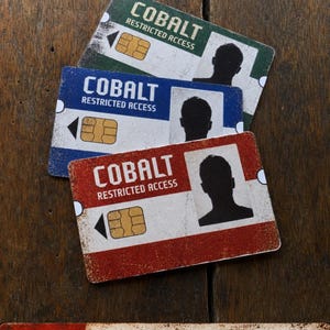 May include: Three rectangular plastic access cards in green, blue, and red, each featuring the text COBALT RESTRICTED ACCESS, a gold chip, and a black silhouette portrait. A separate rectangular sign below reads NFC ENABLED CUSTOM PROGRAMMED FOR YOU.