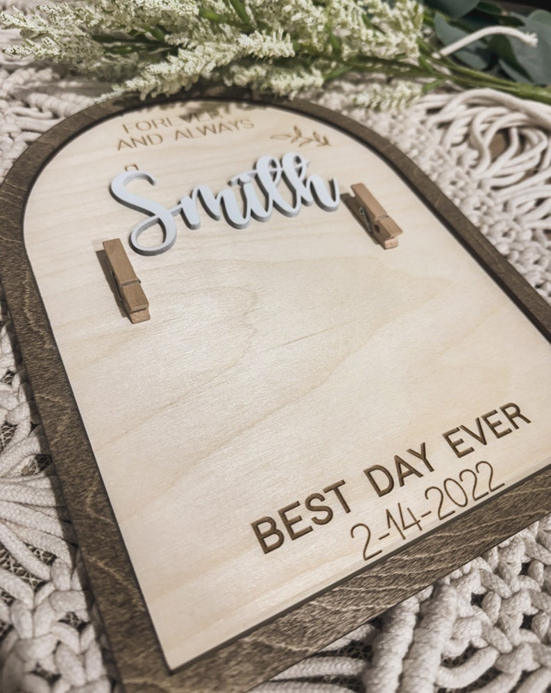 Może przedstawiać: Drewniany znak z brązową ramą i jasnym drewnianym tłem. Znak zawiera tekst "Forever and Always Smith" białymi literami i "Best Day Ever 2-14-2022" brązowymi literami. Na znaku znajdują się dwa drewniane spinacze.