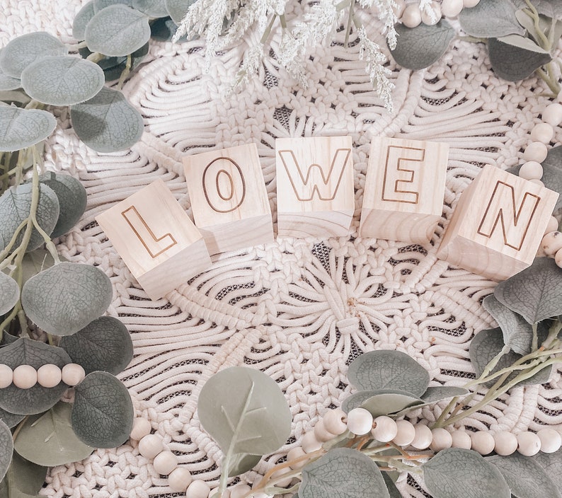 Puede incluir: Bloques de madera que deletrean el nombre "OWEN" en may&uacute;sculas. Los bloques est&aacute;n dispuestos en l&iacute;nea sobre una superficie de crochet blanco con ramas verdes y cuentas blancas.