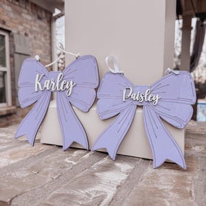 May include: Two purple wooden bow ornaments with the names "Karley" and "Paisley" written in white on them.