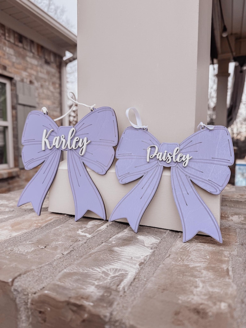 May include: Two purple wooden bow ornaments with the names "Karley" and "Paisley" written in white on them.