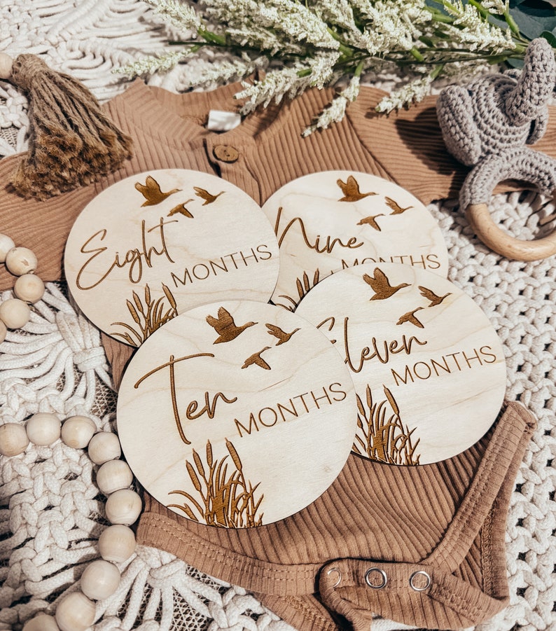 May include: Four wooden baby milestone discs with a rustic brown finish. Each disc features a different month number, the word "months", and a design of flying geese and tall grass. The numbers are eight, nine, ten, and eleven.