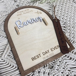 Może przedstawiać: Drewniany łuk z brązową ramą i jasnym drewnianym wnętrzem. Łuk ma wygrawerowane słowa "Forever and Always a Ramirez". Do łuku przymocowane są dwie spinacze do bielizny. Pod łukiem znajduje się znak z napisem "Best Day Ever 09-08-2023". Brązowa sk&oacute;rzana metka z tekstem "Tony Adopted 09-08-2023" wisi na łuku.