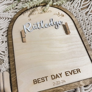Może przedstawiać: Drewniany znak w kształcie łuku z brązową ramą i jasnym drewnianym tłem. Znak głosi "Forever and Always a Rutledge Best Day Ever 2-22-24". Do znaku przymocowane są dwie małe drewniane spinacze.