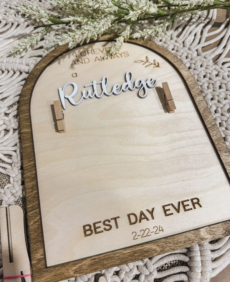 Może przedstawiać: Drewniany znak w kształcie łuku z brązową ramą i jasnym drewnianym tłem. Znak głosi "Forever and Always a Rutledge Best Day Ever 2-22-24". Do znaku przymocowane są dwie małe drewniane spinacze.