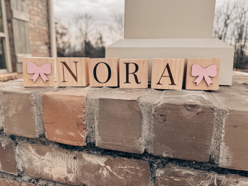 Puede incluir: Bloques de madera que deletrean el nombre "NORA" con lazos rosas en el primer y &uacute;ltimo bloque.