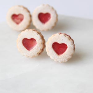 May include: Four heart-shaped cookies with a red filling. The cookies have a light-colored, scalloped edge and a heart-shaped cutout revealing the red filling. The cookies are arranged on a white surface.