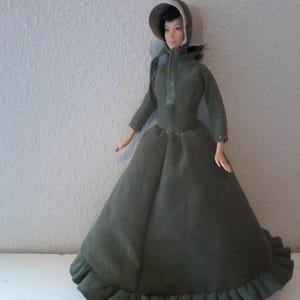 May include: A doll wearing a long, olive green dress with a matching bonnet. The dress has long sleeves and a ruffled hem. The doll has dark hair and is posed against a plain white background.
