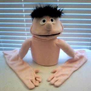 May include: A handmade puppet with a peach-colored head, body, and long arms. The puppet has black hair, large white eyes, and a small nose. The puppet's arms are covered with long peach-colored gloves.