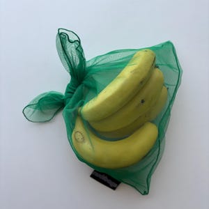 May include: A bunch of ripe yellow bananas inside a green mesh produce bag. The bag is tied at the top with a knot. The bananas are slightly curved and have some brown spots. The bag is on a white background.