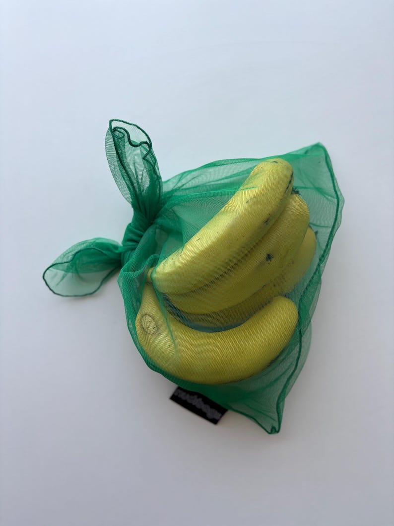 May include: A bunch of ripe yellow bananas inside a green mesh produce bag. The bag is tied at the top with a knot. The bananas are slightly curved and have some brown spots. The bag is on a white background.
