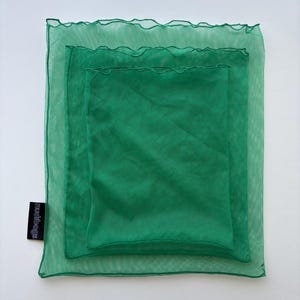 May include: Three nested green mesh bags, each a slightly different shade of green. The bags are square, with a ruffled edge on the largest bag. The smallest bag is solid green. The brand name "mudibags" is printed on a black tag.