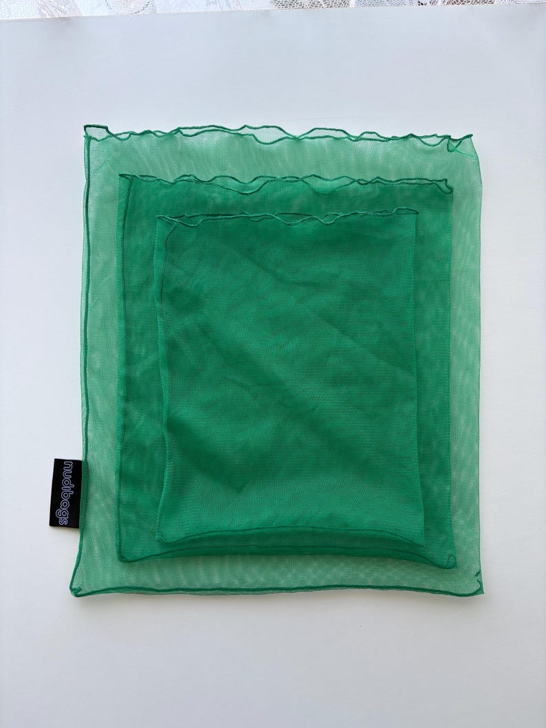 May include: Three nested green mesh bags, each a slightly different shade of green. The bags are square, with a ruffled edge on the largest bag. The smallest bag is solid green. The brand name "mudibags" is printed on a black tag.