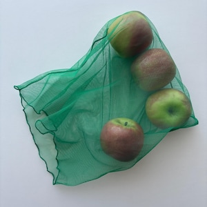 May include: Four apples, some with red and green hues, are held in a green mesh produce bag. The apples are arranged in a cluster, with the bag's fabric draped around them. The background is a plain white surface.