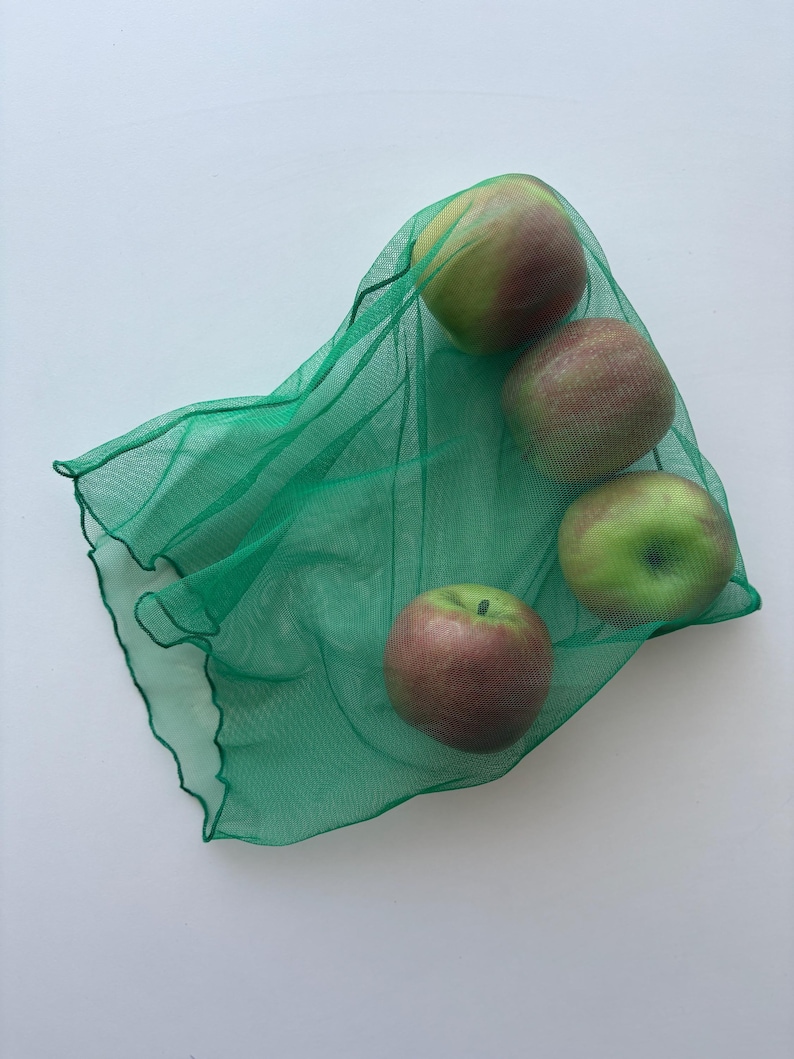 May include: Four apples, some with red and green hues, are held in a green mesh produce bag. The apples are arranged in a cluster, with the bag's fabric draped around them. The background is a plain white surface.