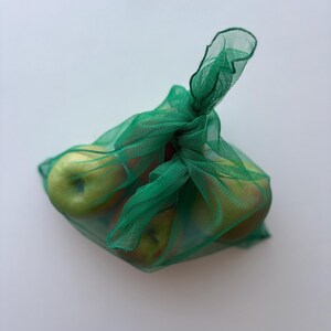 May include: A green mesh produce bag filled with several green apples. The bag is tied at the top with a knot, and the apples are visible through the mesh. The apples have a mix of green and red coloring.