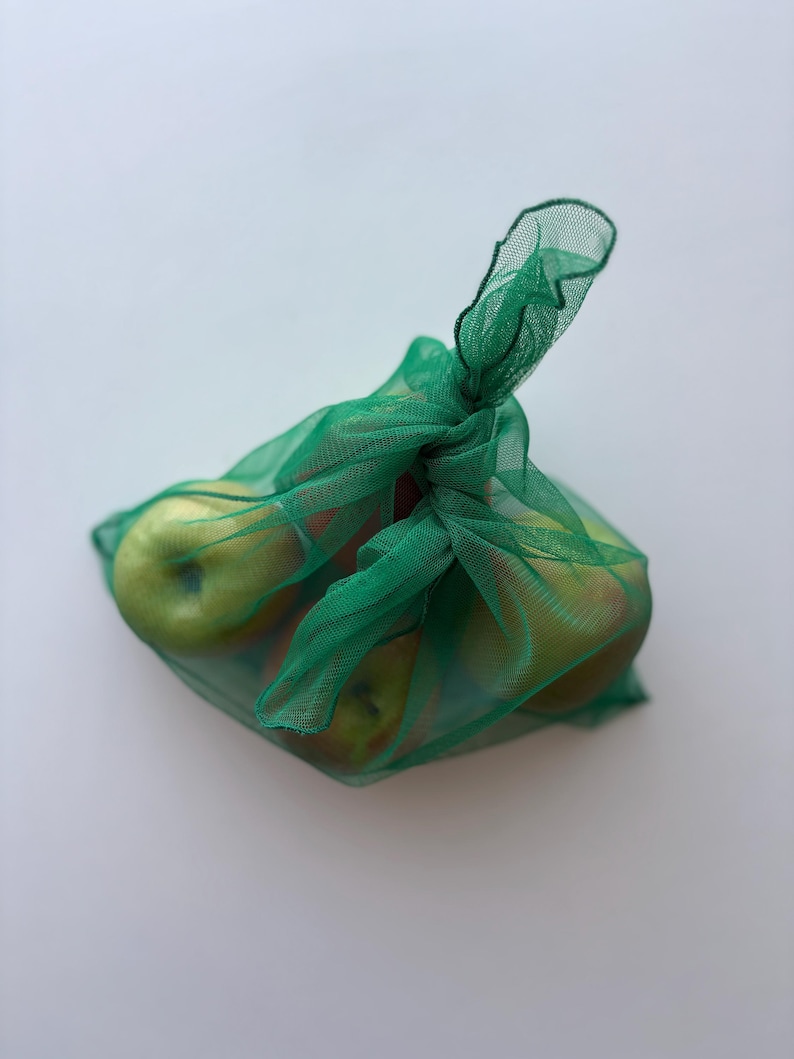 May include: A green mesh produce bag filled with several green apples. The bag is tied at the top with a knot, and the apples are visible through the mesh. The apples have a mix of green and red coloring.