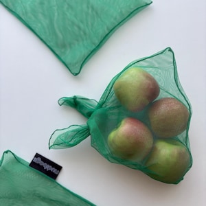 Puede incluir: Bolsas reutilizables de malla verde llenas de manzanas. Las manzanas son rojas y verdes. Las bolsas tienen un cierre de cordón. La etiqueta de la bolsa dice "mudbags".