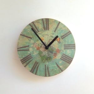 Könnte beinhalten: Eine runde Uhr im Vintage-Stil mit römischen Ziffern und einem floralen Design auf hellblauem Hintergrund. Die Uhr hat schwarze Zeiger und einen Schmetterlingsakzent.