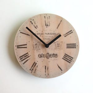 Peut inclure: Une horloge ronde de couleur crème avec des chiffres romains noirs et des aiguilles noires. Le cadran de l'horloge a un aspect vieilli et vintage et présente le texte "Galeries Parisiennes" et "Lille".