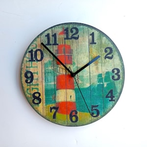 Puede incluir: Un reloj redondo de madera con un diseño de faro. La esfera del reloj tiene un fondo verde y azul con un faro blanco y rayas rojas y blancas. Los números son negros y las manecillas son negras.