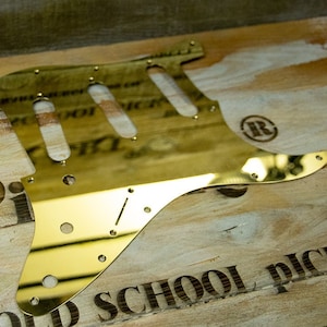 Könnte beinhalten: Ein goldfarbener Gitarren-Pickguard mit einer spiegelnden Oberfläche. Der Pickguard ist gitarrenförmig und hat eine abgerundete Kante. Der Pickguard hat ein Loch für einen Tonabnehmer und ein Loch für eine Buchse. Der Pickguard liegt auf einer Holzoberfläche.