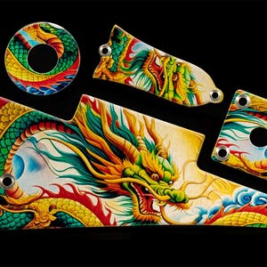 Puede incluir: Un conjunto de protectores de guitarra pintados a mano con un vibrante diseño de dragón. El dragón está representado en tonos verdes, amarillos, rojos y azules sobre un fondo blanco. Los protectores tienen varias formas y tamaños, con el texto "Pick Art".