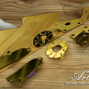 Może przedstawiać: Złote osłony do gitary z misternymi wzorami. Osłony wykonane są z metalu i mają polerowane wykończenie. Osłony są ułożone na drewnianej powierzchni. Pick Art Hand Made Guitars Pickguards.