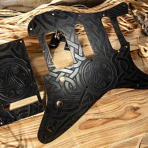 May include: Black guitar pickguard with a Celtic knot design. The pickguard is made of a durable material and features a raised design.