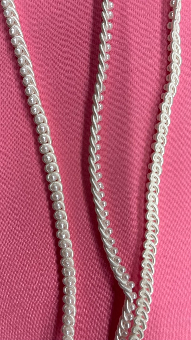 May include: Two white beaded trim cords on a pink background. The cords are made of small white beads and a twisted white fabric.