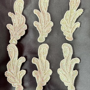 May include: Six decorative leaf-shaped appliques. Each applique is a light green color, covered in small, iridescent sequins. The sequins catch the light, creating a shimmering effect. The appliques are arranged on a dark background.