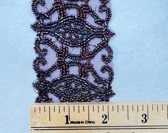 May include: A close-up of a beaded trim with a repeating pattern of swirls and loops. The trim is a dark purple colour with iridescent beads. The trim is measured against a wooden ruler that reads "One Yard".